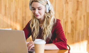 A woman working at home from her laptop, wearing headphones, and drinking coffee.