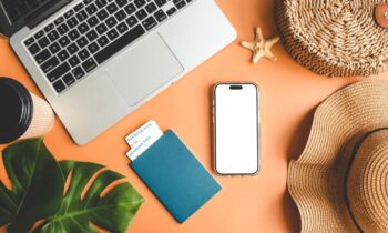 A work from home travel planner's desk, with a laptop, passport, cell phone, and sun hat.