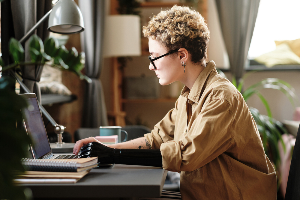A young woman with short hair, glasses, and prosthetic arm working on her laptop reading an article titled side business ideas