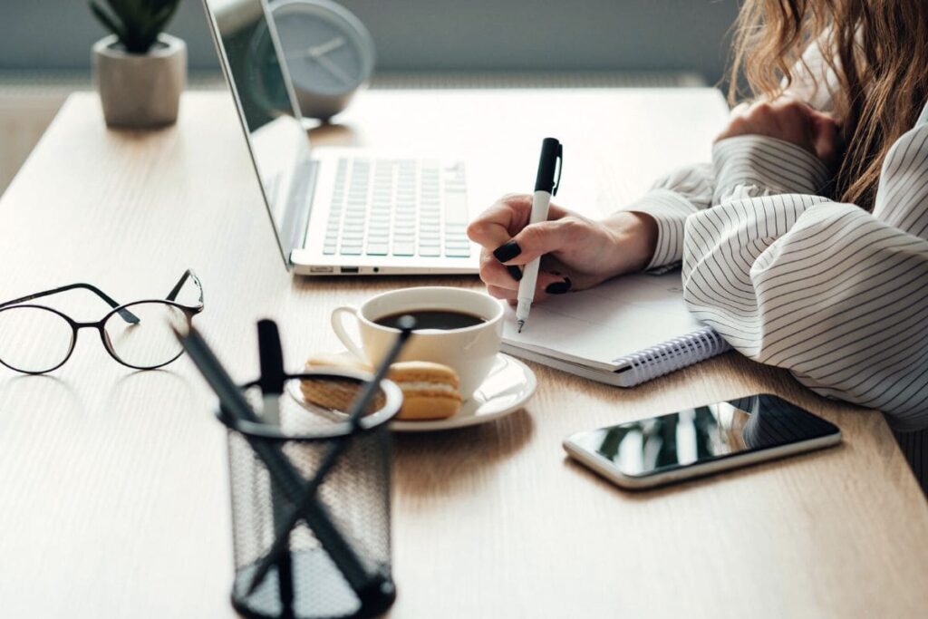 A woman sitting at a desk, working on a laptop and writing in a notebook.