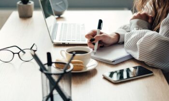 A woman sitting at a desk, working on a laptop and writing in a notebook for blog post find remote work faster.