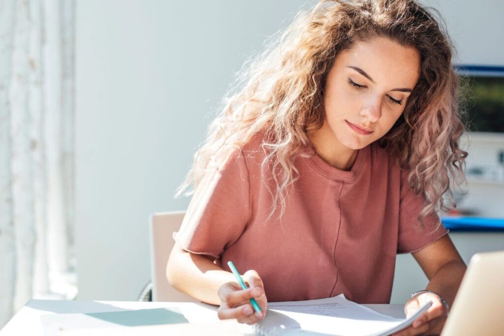 A woman sitting at a desk, working on a laptop and writing in a notebook.