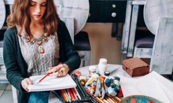 A woman sitting outdoors at a table and using colored pencils to draw in a large sketchbook.