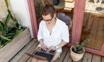 Woman sitting on a wooden patio next to potted plants, using a laptop to job hunt for remote jobs.