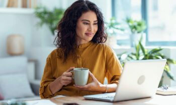 A woman working from her home office desk, holding a coffee cup, smiling, and reading on her laptop for blog post get promoted at work.