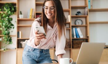 A work at home business woman standing at a desk in her home office, looking at something on her phone for blog posy make money on Pinterest