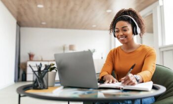 A freelancer working remotely from her kitchen table, using a laptop, wearing headphones, and writing in a notebook for blog post sell services online