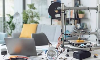 A home office desk, with a laptop, 3D printer, and other fabricating tools for blog post small machines that make money.