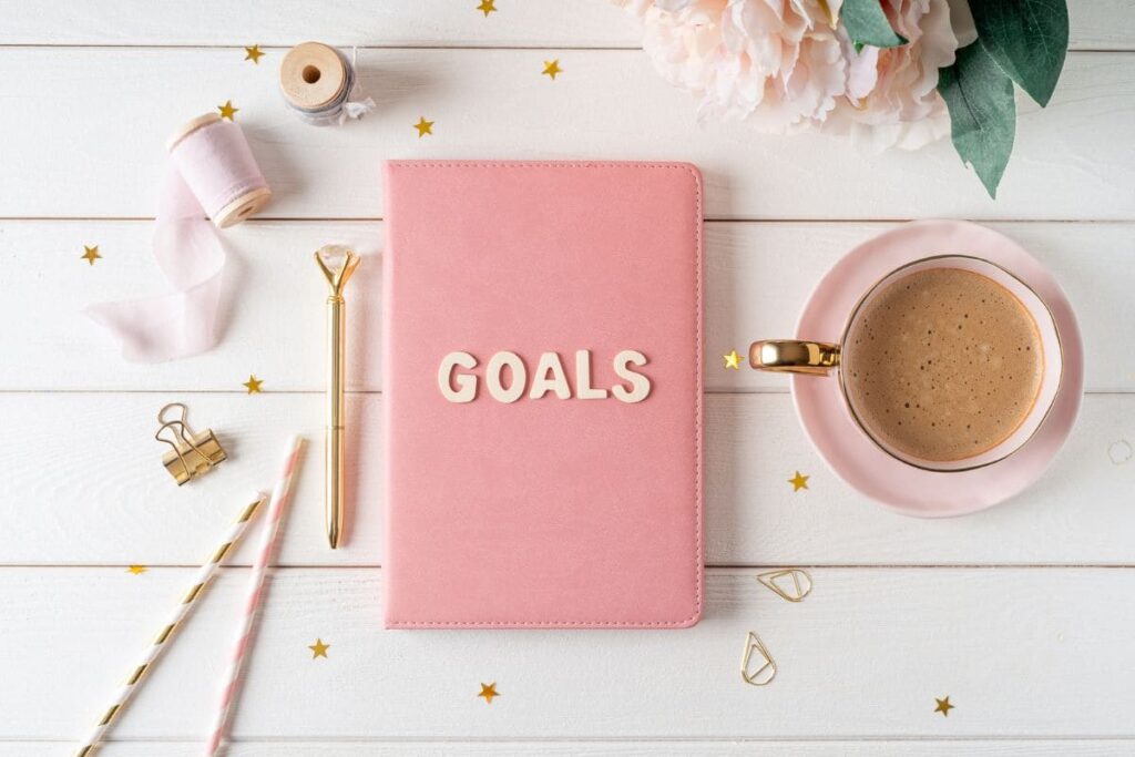 A notebook labeled, "Goals" sitting on a woman's desk, with coffee and flowers.